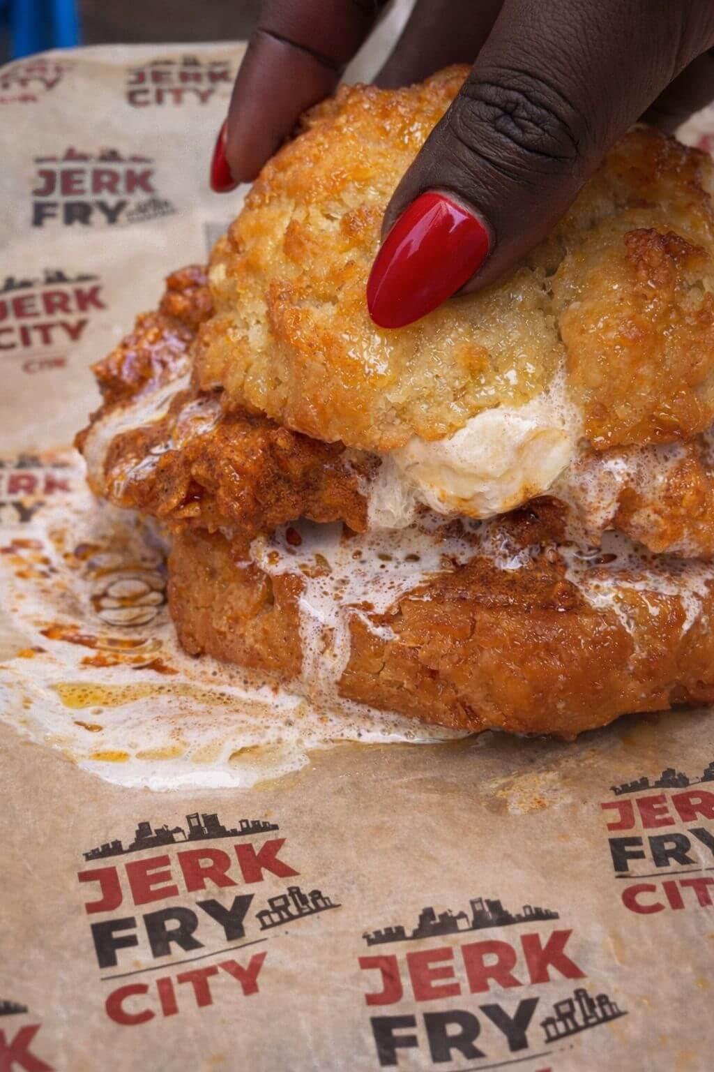 Fried chicken sandwich with a biscuit on top, held by a hand with red nail polish, on a 'Jerk Fry City' branded paper - Jerk Fry City