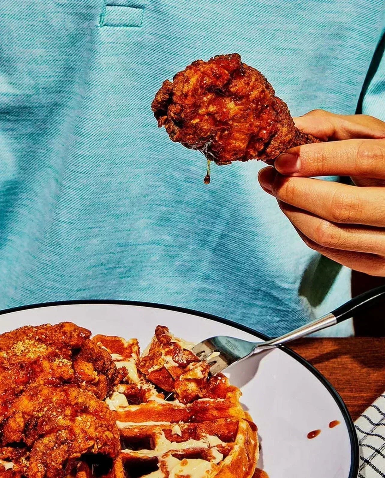 Fried chicken being held above a plate of waffles with a blue background - Jerk Fry City