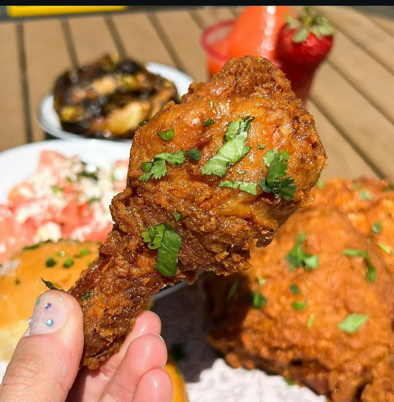 Fried chicken with green herbs held by a hand, with a blurred background of food and a straw - Jerk Fry City