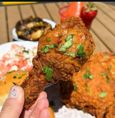 Fried chicken with green herbs held by a hand, with a blurred background of food and a straw - Jerk Fry City