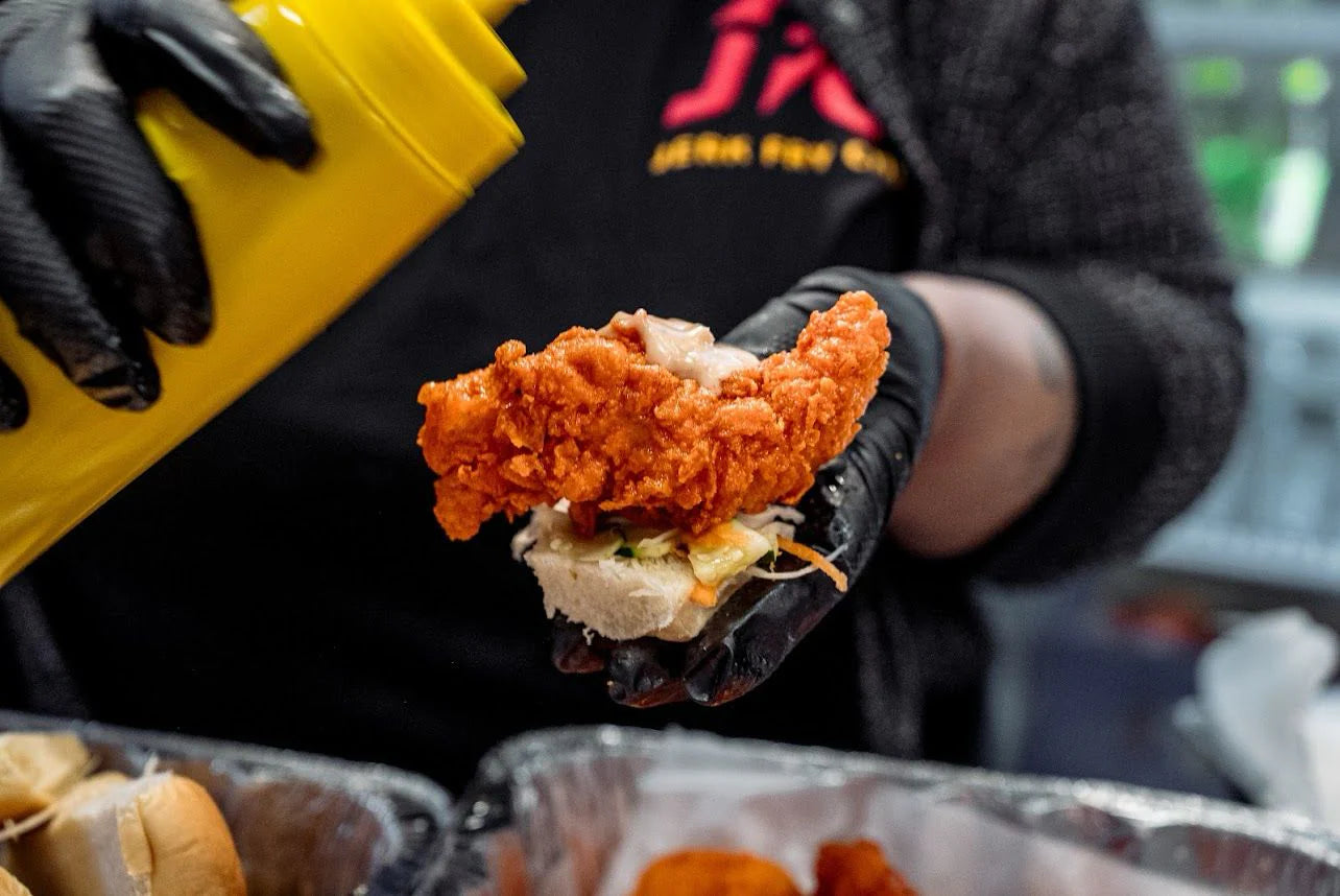 Person holding a sandwich with fried chicken and cheese, wearing black gloves and a black jacket with red text.