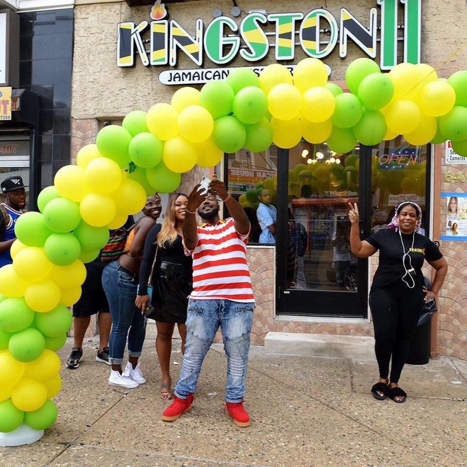 People posing under a colorful balloon arch in front of a Jamaican restaurant.