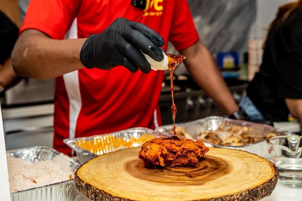 Person in red shirt and black gloves pouring sauce over fried chicken on a wooden board - Jerk Fry City